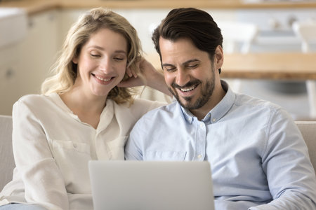 Pretty millennial 35s couple laugh while watch movie use TV streaming services, choose goods, buy travel tour, booking hotels planning joint holiday, enjoy carefree weekend time on internet use laptopの写真素材
