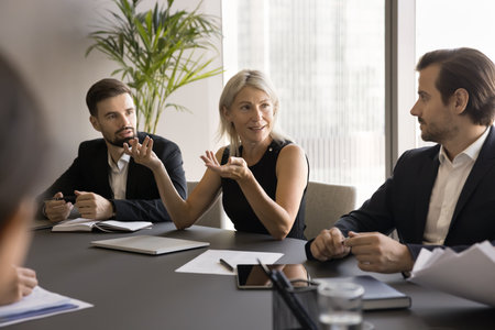 Positive confident mature business woman speaking to younger project partners on corporate meeting,...
