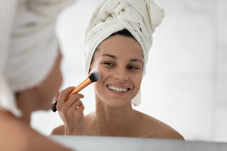 Happy millennial girl look in mirror in bathroom use cosmetics brush put powder or beauty product on face cheeks, smiling young woman get ready do daily make up in morning in bath after showerの写真素材