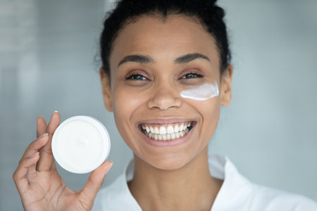 Portrait of happy african American girl use moisturizing face cream, recommend beauty product for healthy glowing smooth skin, smiling biracial woman do morning facial procedures, skincare conceptの写真素材