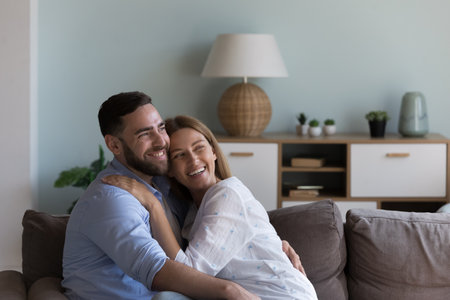 Happy attractive married couple hugging at home with love, sitting on couch, looking away, thinking, dreaming, planning, laughing, smiling, celebrating anniversary, family eventの写真素材