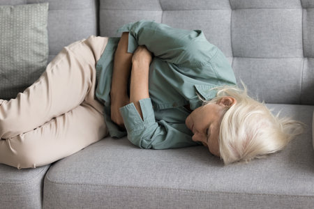 Upset exhausted older woman suffering from strong pain, health problems, serious disease symptoms, feeling stomach ache, resting on home couch with arms folded on bellyの写真素材