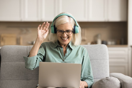 Positive elderly student woman in wireless headphones using laptop on sofa, saying hello at webcam, waving greeting hand, speaking on online video conference call at homeの写真素材