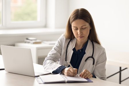 Female general practitioner in white coat sits at desk in hospital writes notes in paper notepad, work use laptop. Doctor manages appointment, fill patient anamnesis or client information in journalの写真素材