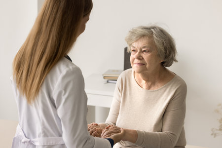 Older woman visit to doctor for medical checkup in hospital. Caring young therapist in white uniform holding hands of patient inform good news about treatment, support encouraging old person. Medicareの写真素材