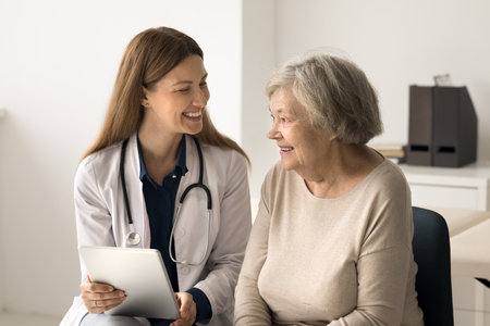 Smiling young female doctor in uniform hold digital tablet shows result of medical screening examination, give consultation, explain electronic prescription. Older patient receiving professional helpの写真素材