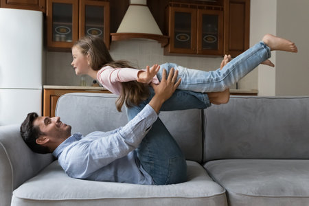 Sportive strong dad keeping balance with happy little kid girl on legs, resting on sofa, holding, lifting child in air, doing acroyoga at home. Daughter playing airplane flight with daddyの写真素材