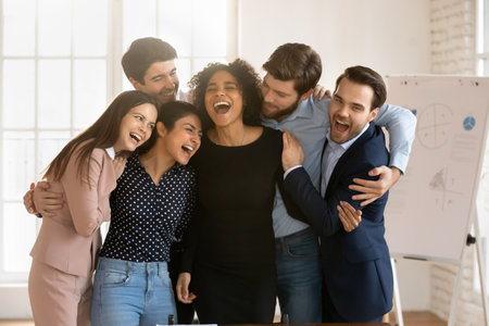 Cheerful excited multiethnic office friends celebrating teamwork success, cooperation, congratulating on young African business team leader on successful project, hugging, laughingの写真素材