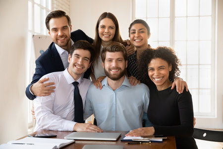 Positive young diverse business colleagues meeting at workplace table, standing close, hugging with joy, looking at camera. smiling, laughing. Successful united team portraitの写真素材