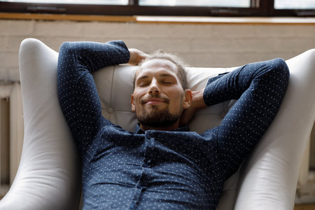Happy peaceful sleepy young millennial man in casual relaxing with closed eyes at home, taking deep...