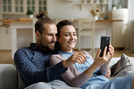 Happy sweet young couple taking selfie picture on mobile phone, watching live media content, talking on video call, using online app on digital gadget, relaxing on couch, enjoying leisure at homeの写真素材