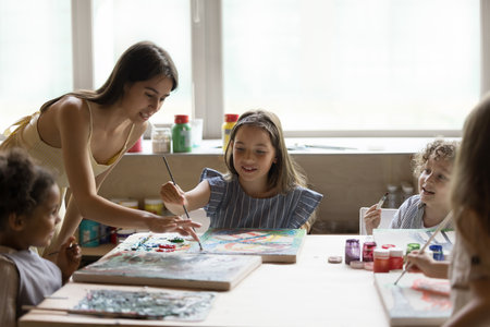 Group of little multiethnic boys and girls sit at table painting on canvas attend creative art-class led by woman painter, teacher helps, teach to preschoolers in art-studio, talent development, hobbyの写真素材