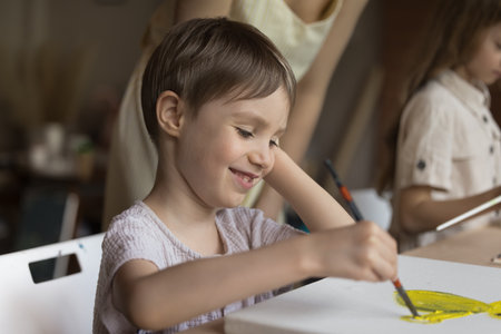 Cute little boy sit at table holding paintbrush makes yellow paint strokes on canvas, smile, enjoy painting group class, close up. Kids development, hobby to improve creative ability, self-expressionの写真素材