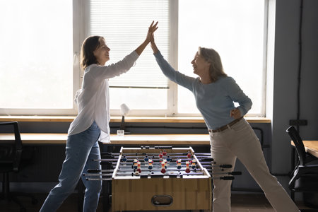 Young and mature women, two office employees playing table football, give high five, celebrate...