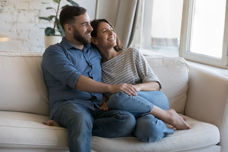 Happy dating couple enjoying relaxation, leisure time at cozy home, discussing romantic plans, dreaming together, looking at window away, holding hands, hugging with love, affectionの写真素材