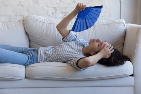 Exhausted sick homeowner woman resting on home sofa, waving handheld paper fan, suffering from fatigue, hypoxia, headache, heating stroke, holding head, swiping sweatの写真素材