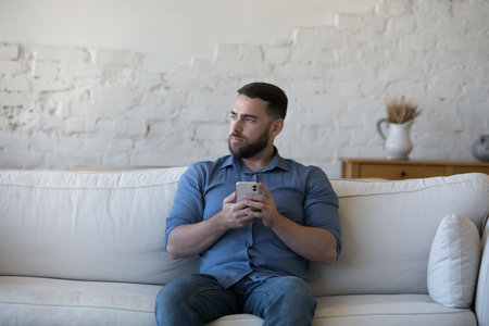 Serious thoughtful mobile phone user man holding gadget, sitting on sofa, looking away, thinking over problems, bad news, making decision, choice, dreaming, ponderingの写真素材