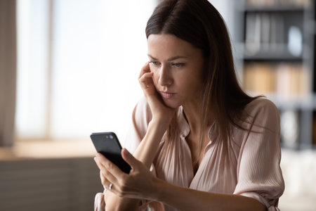 Upset woman feels concerned about received message holds smartphone looks at screen reading sms sitting in living room alone, close up view. E-banking statement overcharging, negative bad news conceptの写真素材