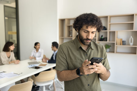 Focused young Arab business professional man typing on mobile phone while team on coworkers talking...