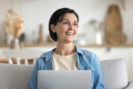 Cheerful mature woman spend time at home with laptop, enjoy e-shopping, purchasing on-line, use marketplace e-commerce services, chatting with friend in social media, having fun on weekend on internetの写真素材