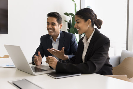 Couple of happy Indian business coworkers talking on video call at laptop together, sitting at computer, watching online content, smiling, laughing, cooperating on Internet projectの写真素材