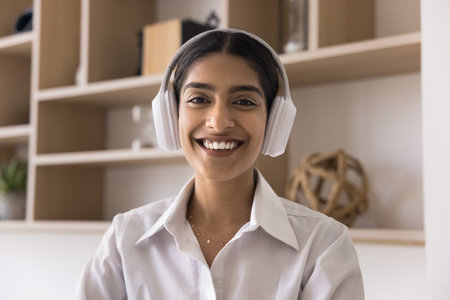 Cheerful pretty Indian manager girl in headphones looking at camera, smiling, speaking on video...