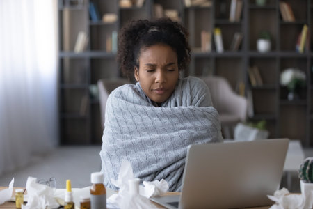 Frustrated ill young Black woman, remote employee, patient wrapped in warm plaid suffering from cold, fever, flu, influenza, taking pills, making video call, consulting doctor online, using laptopの写真素材