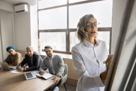 Serious pretty mature female business leader writing on whiteboard on meeting with multiethnic team, explaining project plan, strategy, sharing ideas with office colleagues, giving report, seminarの写真素材