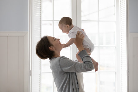 Happy young mother play with newborn at home, standing in cozy light domestic room, lifts, raise up...