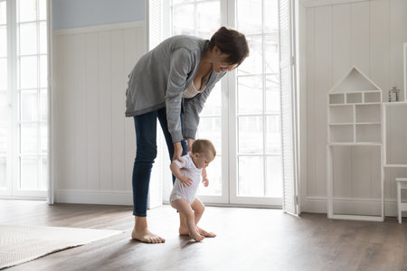 Cute toddler in bodysuit holding loving mothers hands and makes first steps in cozy bedroom, walk on...