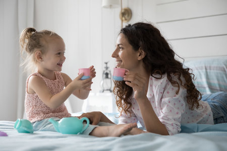 Mother lying on bed in bedroom spend time with cute little daughter, play together, drink morning tea from plastic toy cups, enjoy carefree girlish talk, enjoying funny leisure and playtime at homeの写真素材