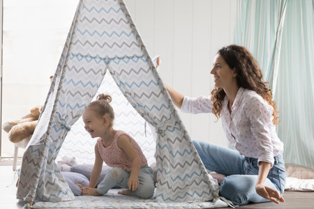 Happy beautiful mother enjoy playtime in modern, warm playroom with cute little daughter, playing together in toy tent seated on floor, having fun, spend leisure at cozy home. Playtime, family gamesの写真素材