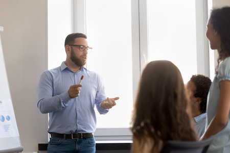 Serious confident busy project leader talking to employees on team meeting. Business coach, mentor teaching students, interns, explaining work tasks. Businessman presenting project to colleaguesの写真素材