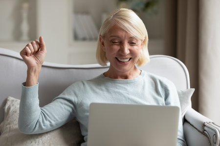 Excited mature woman using laptop, reading message, celebrating online success, good news receiving,...