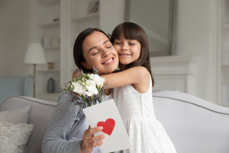 Little daughter hugging happy young mother, congratulating with birthday or mothers day, smiling mother holding flowers and greeting card with red heart, sitting together on cozy sofa at homeの写真素材