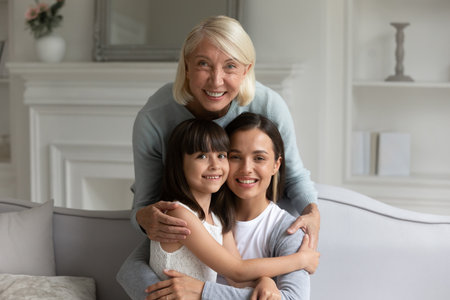 Head shot smiling three generations of women at home, happy older grandmother hugging granddaughter...