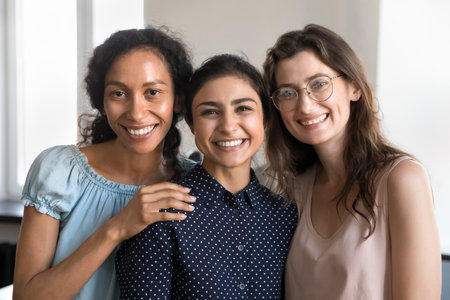 Diverse business team of happy young employees women standing close, looking at camera, smiling, laughing, hugging. Female colleagues enjoying cooperation, teamwork result, office friendshipの写真素材