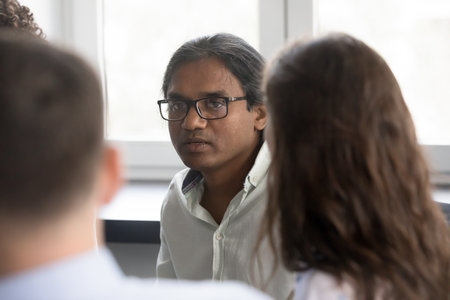 Serious engaged Indian team employee in glasses talking to coworkers, sharing idea, offering solution, brainstorming, discussing task. Addict man speaking on group therapy meetingの写真素材