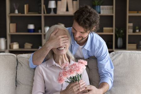 Millennial caring son covering mature mothers eyes with palm, gives her carnation flowers, makes pleasant surprise during his visit at mommy home. 8-march or life event celebration, attention, loveの写真素材