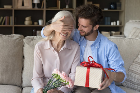 Loving grown up adult son covering smiling mature mothers eyes with palm, hold gift box, makes pleasant surprise on birthday, celebrate anniversary, family holiday at home. Life event, attention, loveの写真素材
