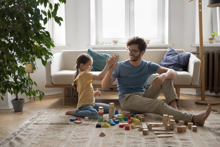 Handsome father and little daughter spend leisure time together sitting on floor in living room. Young dad enjoy friendly communication with child, play with wooden blocks, giving high five look happyの写真素材