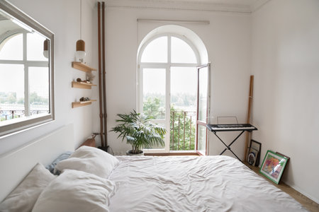 Cozy light bedroom with big mirror on wall, shelves, synthesizer, white wrinkled linen on double bed, view to summer city through opened door, no people. Interior of stylish, cosy modern empty bedroomの写真素材