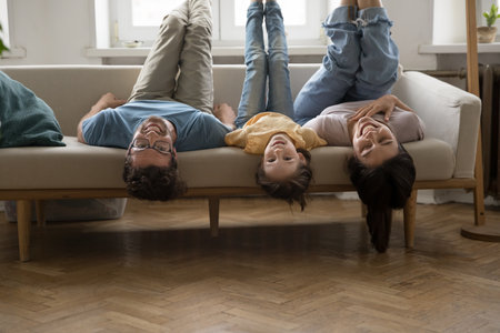 Family fooling around lying on sofa upside down, laughing and looking at camera. Carefree active parents with little daughter having fun, exercising together with legs raised on cozy couch. Leisureの写真素材