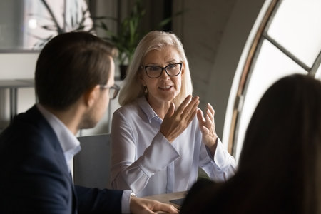 Engaged confident senior business leader woman speaking to group of colleagues on brainstorming meeting, discussing teamwork, cooperation. Mature boss talking to managers, offering work decisionの写真素材