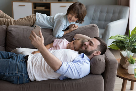 Happy relaxed millennial dad and cheerful little sibling kids lying, hugging on sofa, using online app on mobile phone, taking selfie photo, talking on video call, smiling, laughingの写真素材