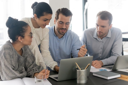 Multiethnic employees gather at desk in office work together on laptop discussing company project....