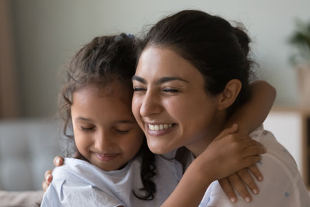 Lovely young Indian woman enjoy embraces of preschooler 6s pretty daughter sit on sofa at home....