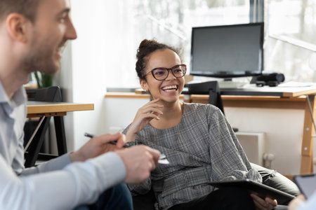 Smiling young African American woman employee have fun talking brainstorming with diverse colleagues at meeting in office. Happy ethnic female worker cooperate with coworkers at briefing at workplace.の写真素材
