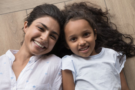 Young beautiful Indian mother and little preschooler 6s daughter lying together on warm floor smile staring at camera feel happy, close up shot. Family portrait, heredity, offspring, bonding conceptの写真素材