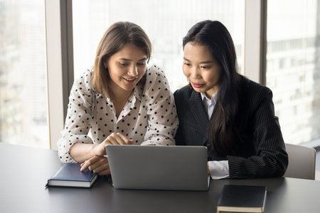Two positive diverse young colleague girls talking at workplace, working on project together laptop, meeting for teamwork, collaboration, discussing online startup strategyの写真素材